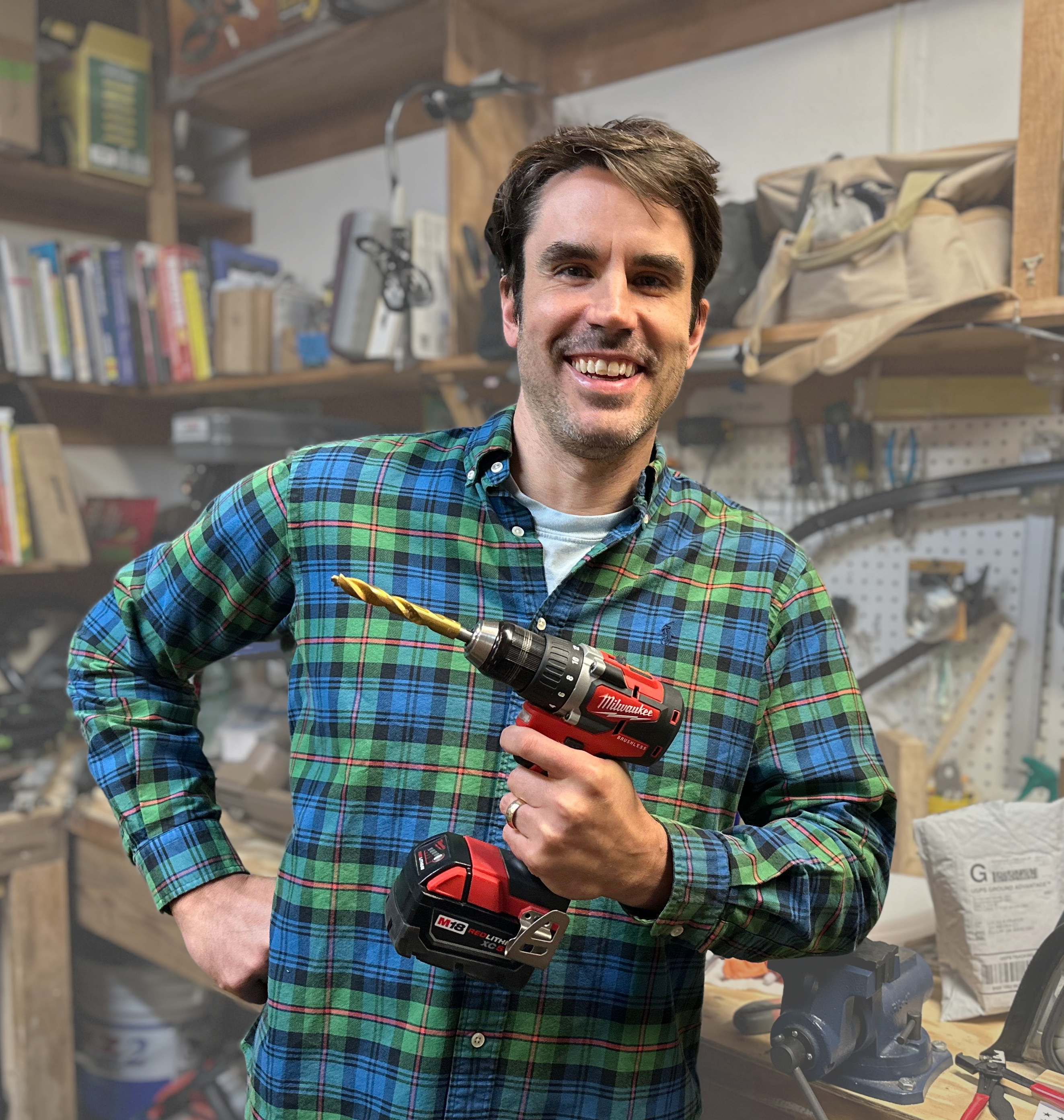 Luke Prater smiling in his workshop, holding a power drill in front of bookshelves and a pegboard of tools